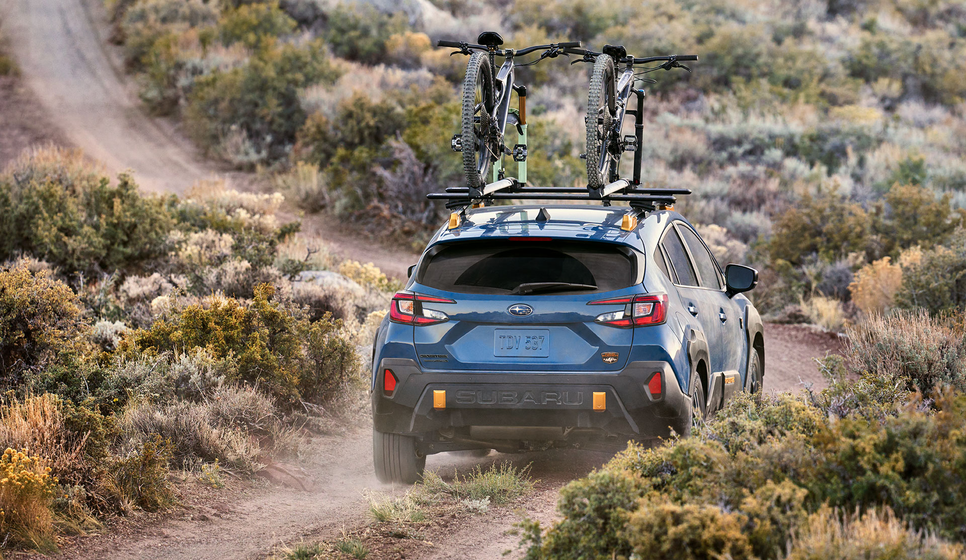 Rear shot of 2024 Crosstrek Wilderness driving with bicycles on roof racks along dusty trail.